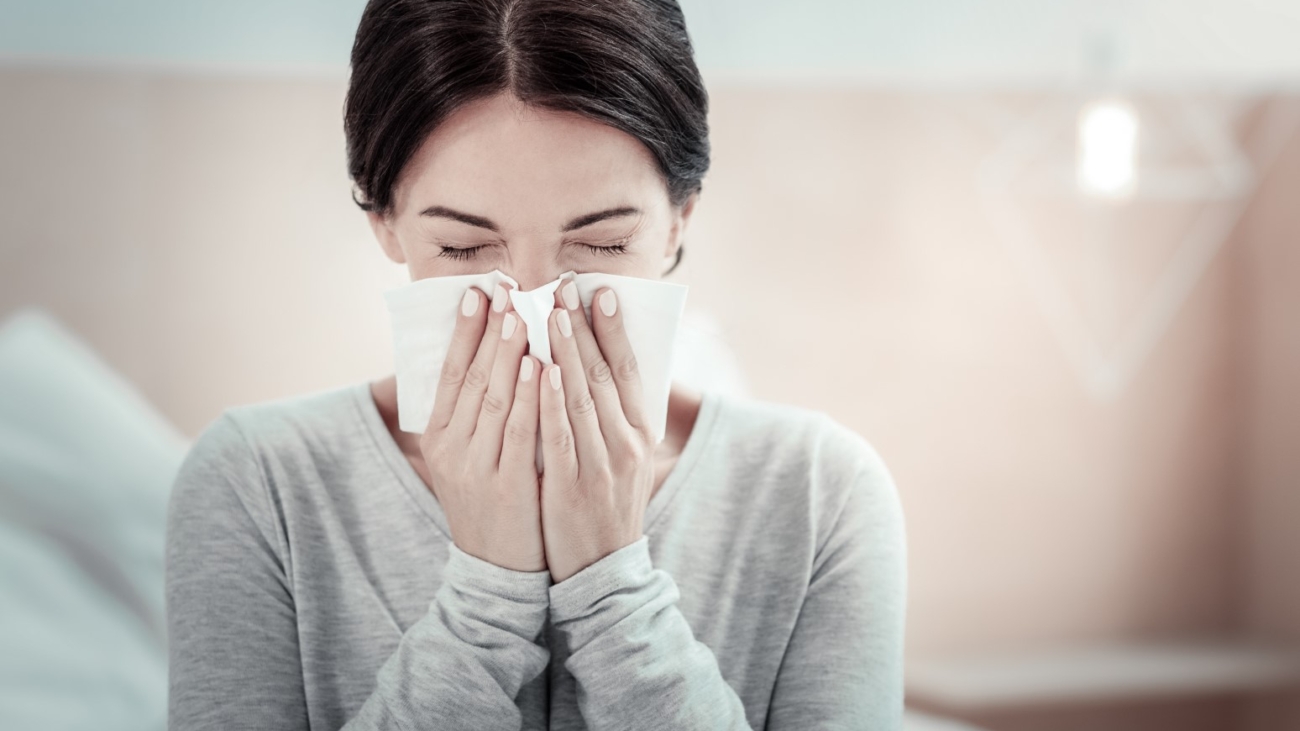 Home treatment. Ill exhausted unhappy woman sitting in the bright room holding a napkin and sneezing.
