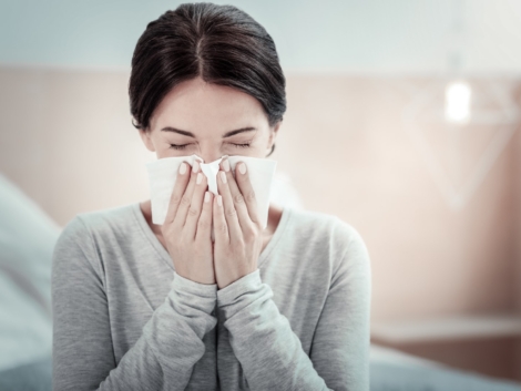Home treatment. Ill exhausted unhappy woman sitting in the bright room holding a napkin and sneezing.
