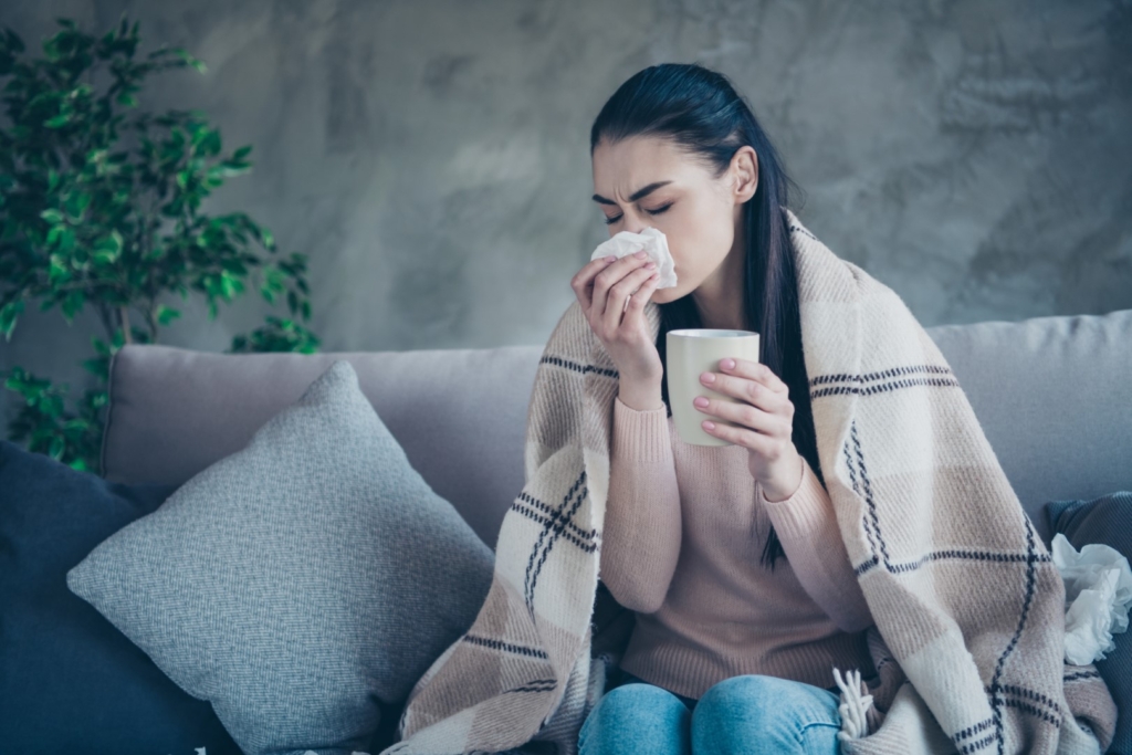 Photo of suffering lady with cup and napkin caught cold drinking medicine from flu covered with blanket, wearing pullover and jeans sitting sofa