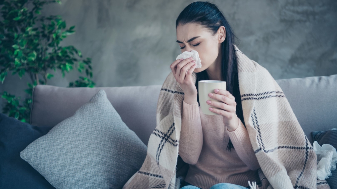 Photo of suffering lady with cup and napkin caught cold drinking medicine from flu covered with blanket, wearing pullover and jeans sitting sofa