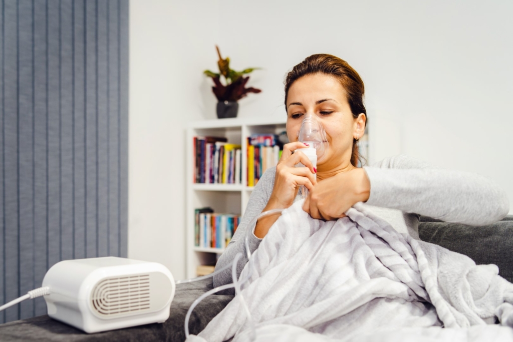 Woman lying covered with blanket using vapor steam inhaler nebulizer mask inhalation at home on the bed medicament treatment asthma pneumonia bronchitis coughing sick