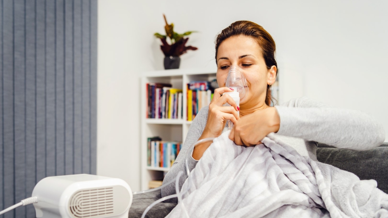 Woman lying covered with blanket using vapor steam inhaler nebulizer mask inhalation at home on the bed medicament treatment asthma pneumonia bronchitis coughing sick