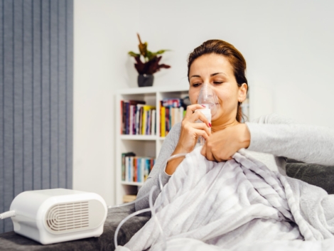 Woman lying covered with blanket using vapor steam inhaler nebulizer mask inhalation at home on the bed medicament treatment asthma pneumonia bronchitis coughing sick