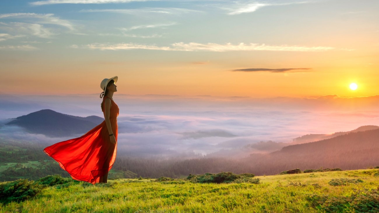 Young woman in red long dress standing in morning mountains with rising sun and white fog below enjoying view of idyllic nature.