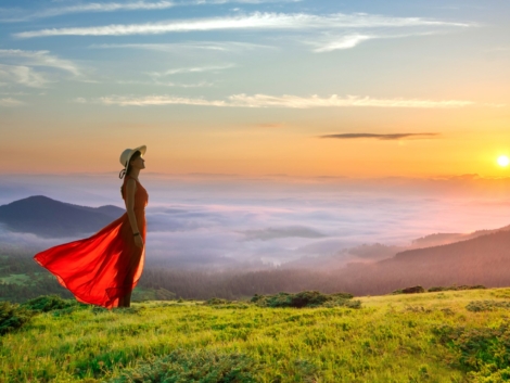 Young woman in red long dress standing in morning mountains with rising sun and white fog below enjoying view of idyllic nature.
