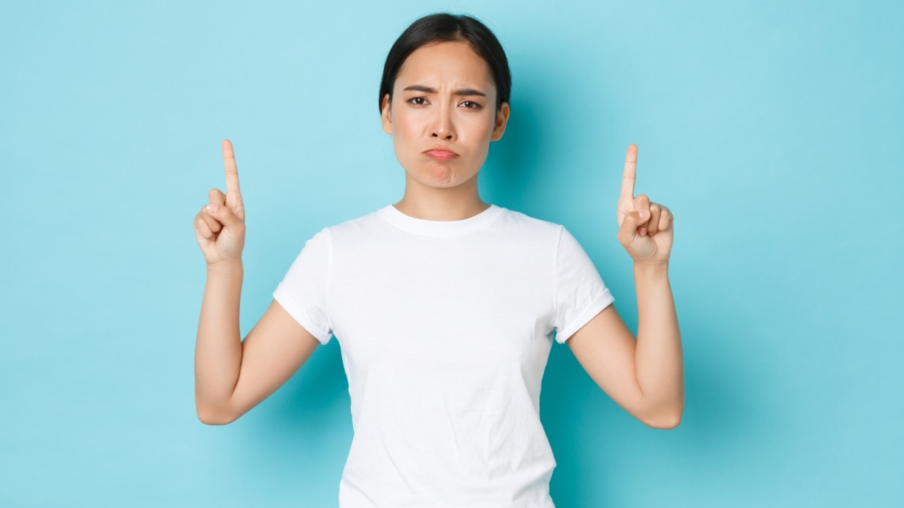 Upset and bothered asian girl in casual outfit, sulking disappointed, pointing fingers up displeased, complaining over bad quality, awful promo banner, standing blue background.