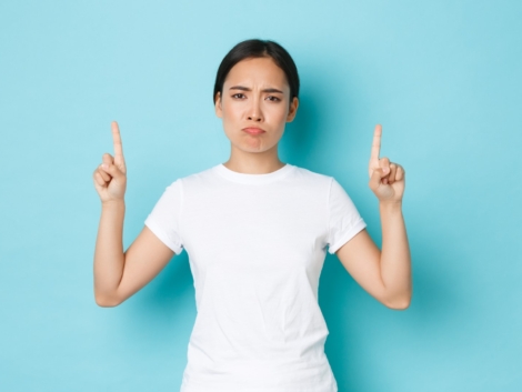 Upset and bothered asian girl in casual outfit, sulking disappointed, pointing fingers up displeased, complaining over bad quality, awful promo banner, standing blue background.