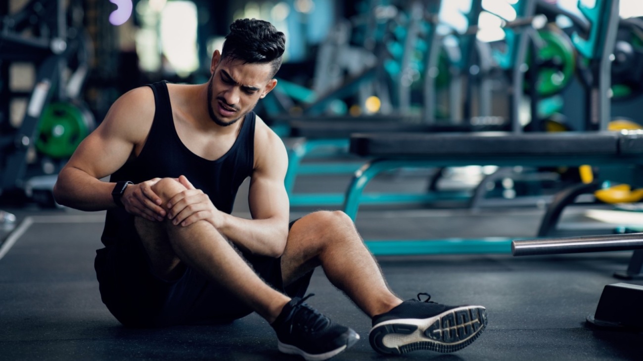 Sport Trauma. Young Arab Male Athlete Touching His Hurted Knee At Gym, Upset Middle Eastern Guy Sitting On Floor And Massaging Sore Injured Leg After Workout Training In Fitness Club, Free Space