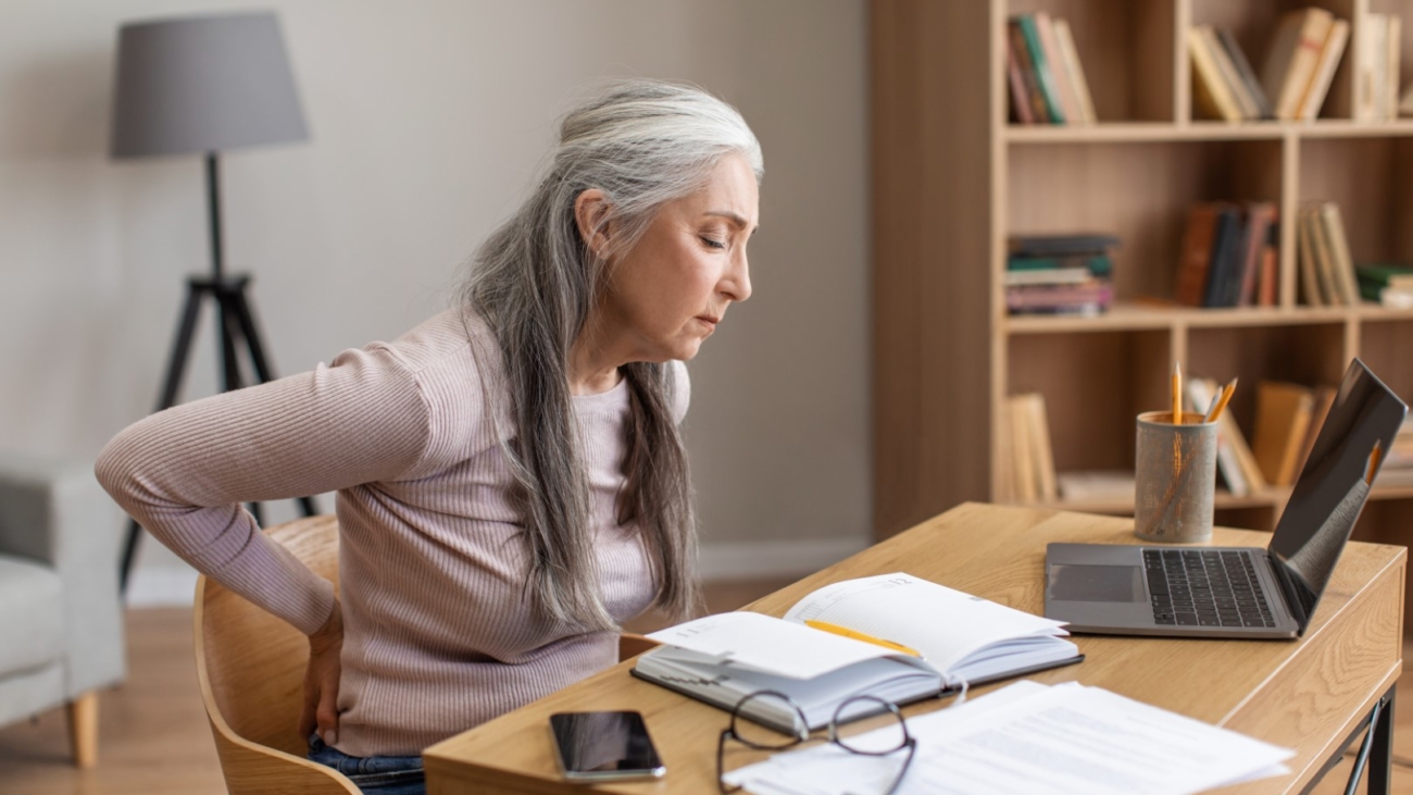 Unhappy caucasian old female with gray hair works on computer and suffers from back pain in room interior. Overwork, arthritis and health problems, study and business remotely at home during covid-19