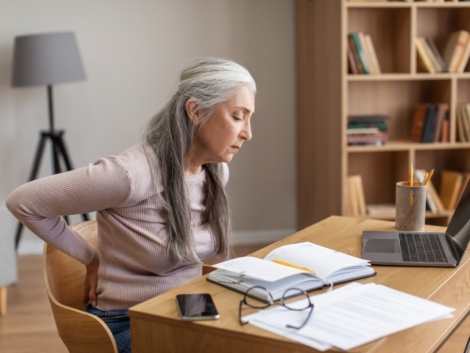 Unhappy caucasian old female with gray hair works on computer and suffers from back pain in room interior. Overwork, arthritis and health problems, study and business remotely at home during covid-19